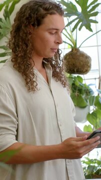Vertical video: Raising phone, person checking messages at window wearing beige shirt, kokedama