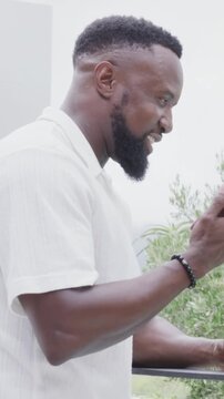 Vertical video: Laughing man in white shirt leaning on balcony railing, seeing phone and scrolling