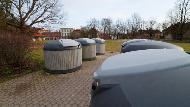 Row of underground municipal waste dumpsters in an urban park on a paved walkway, with leafless trees and overcast sky&mdash;public sanitation and neighborhood trash collection.