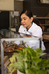 Fototapeta premium Woman pastry chef wearing white jacket with black trim arranging pastries in glass display case