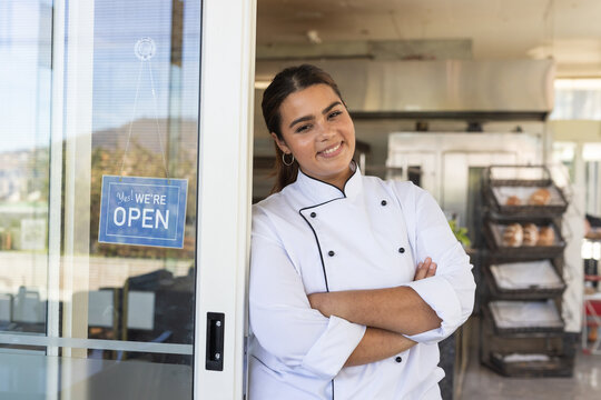 Female chef standing at bakery door wearing white jacket, leaning with arms crossed, racks visible