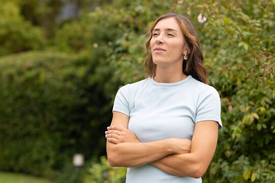 Woman standing with arms crossed, gazing upward in garden, wearing light blue top and earrings