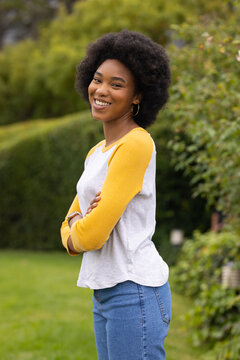 African woman standing arms crossed smiling wearing raglan shirt jeans hoops wristwatch in garden