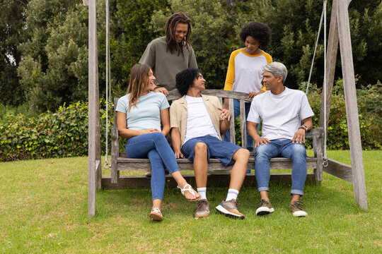 Diverse friends sitting on wooden swing bench in backyard, chatting while wearing casual clothing