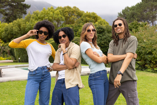 Diverse friends posing on lawn by pool, wearing sunglasses and smartwatch with casual clothing