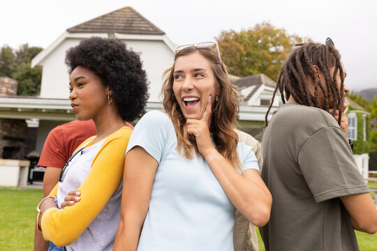 Diverse friends posing back-to-back on front lawn at gathering, woman touching face with sunglasses