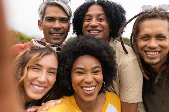Diverse friends smiling, leaning close for selfie at park with smartphone and sunglasses