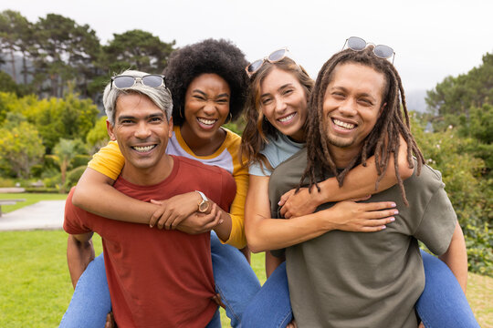 Diverse friends posing piggyback on park lawn, wearing sunglasses, jeans, wristwatch, colorful tees