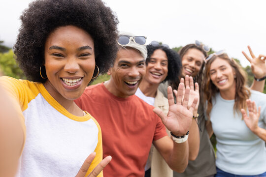 Diverse friends posing for selfie in park, African American woman holding smartphone and waving