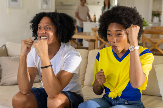 African American man and woman watching live match on TV from sofa, wearing jersey, pumping fists