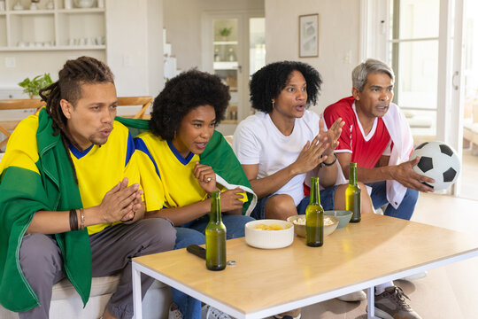 African American friends watching soccer match on sofa at home holding soccer ball, wearing jerseys