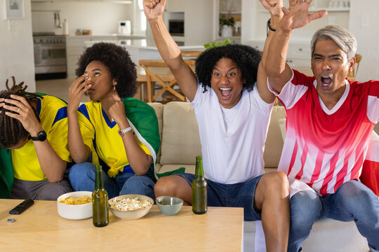 Diverse group wearing soccer jerseys watching match at home, cheering by coffee table with beer