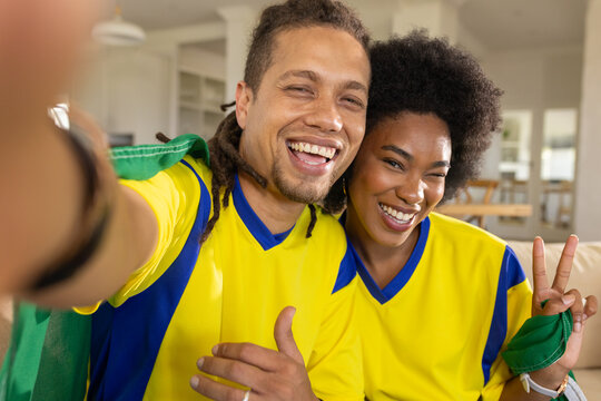 African American couple sitting on sofa wearing yellow-blue kits, taking selfie holding green scarf