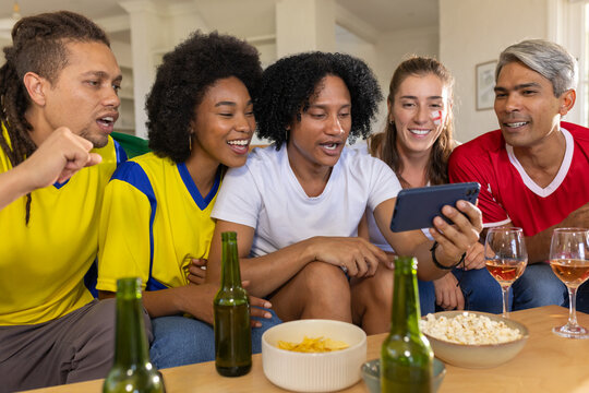 Diverse friends wearing jerseys leaning forward watching smartphone on sofa with snacks and drinks