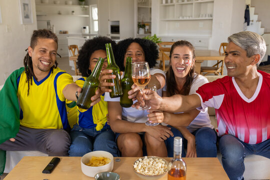 Diverse friends clinking beer bottles and wine glass while sitting on couch wearing sports jerseys