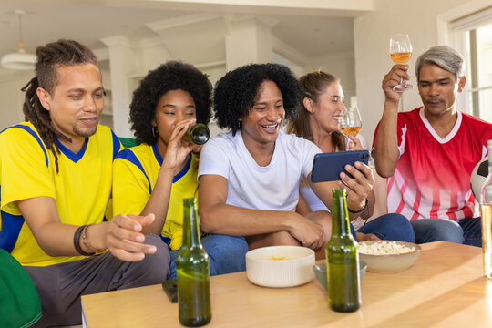Diverse group in sports jerseys raising wine glasses and toasting while watching smartphone on sofa