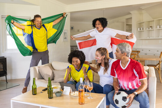 Diverse friends wearing jerseys cheering and watching soccer on tablet with flags and soccer ball