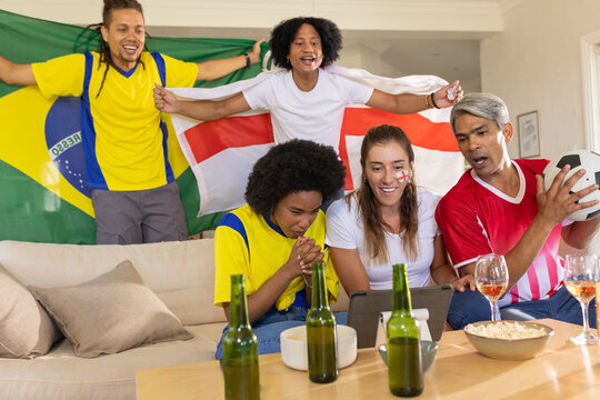 Diverse friends wearing jerseys watching soccer on tablet at home holding Brazil and England flags