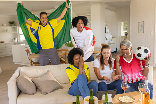 Diverse friends in jerseys cheering and waving flags while watching match on tablet in living room