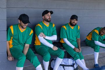 Diverse male teammates sitting on bench in block dugout wearing green jerseys, duffel at feet © wavebreak3