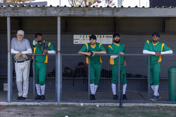 Diverse male baseball team and coach standing on dugout rail in green uniforms holding gloves, bat © wavebreak3