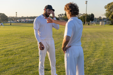Male cricket players standing in white uniforms on grass, one holding red ball while other pointing © wavebreak3