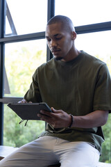 African American man sitting by metal window holding tablet in case wearing olive green T-shirt © wavebreak3