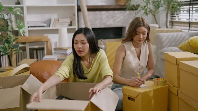 Moving day relocation in apartment women friends packing cardboard box with tape at home, busy young roommates organizing living room for moving and packing cardboard boxes for home relocation