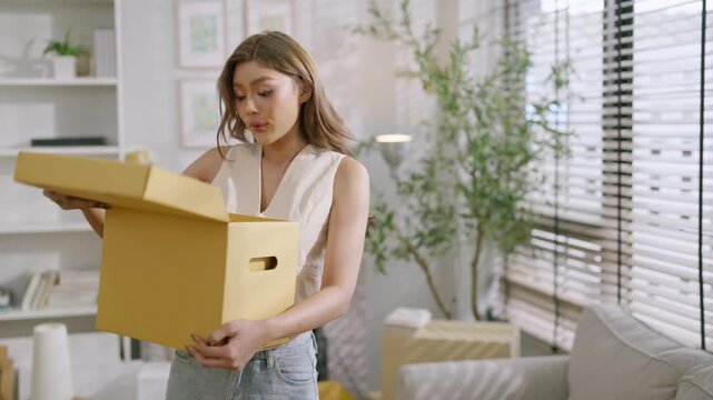 Young woman with cardboard box moving and unpacking in new home apartment living room, relocation day in bright home interior with daylight, settling in and celebrating delivery during unpacking