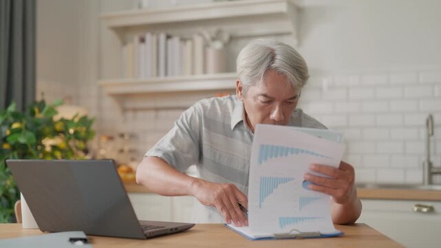 Senior man in home office at kitchen table reviewing paperwork and document on laptop for finance budget accounting, reading financial report, analyzing records with focused expression