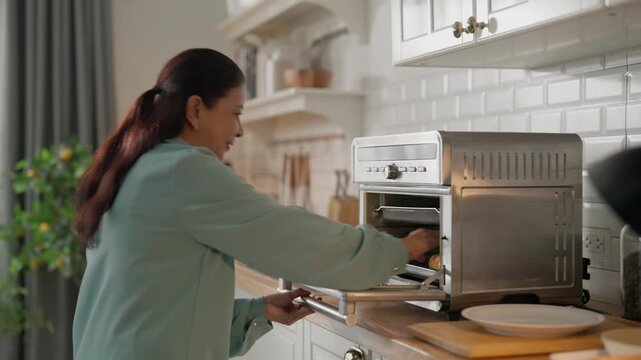 Family kitchen candid indoors conversation home, adult woman back view using toaster oven baking breakfast pastry, adjusting control knob while talking with senior man, casual morning cooking routine