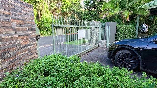 Automatic metal security gate opening for black electric car entering luxury residence driveway.