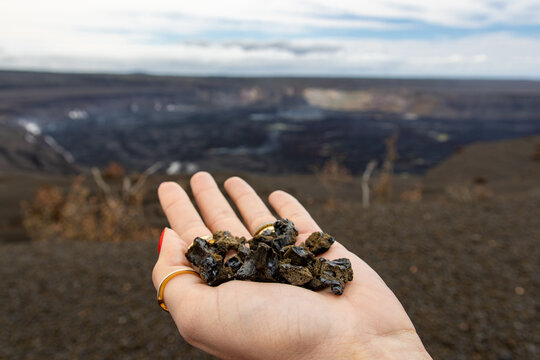 Person holding volcanic lapilli cinders in their hand with the Kilauea Crater in the background on the Big Island of Hawaii