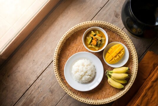 Top-down flat lay of a monk's simple morning meal arranged on a traditional Thai offering tray &mdash; rice, curry, fruit &mdash; with edge of saffron robe and alms bowl visible, warm natural window light.