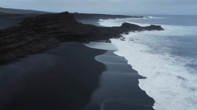 Waves crash on rugged shoreline with black sand beach and rocky terrain under cloudy skies.