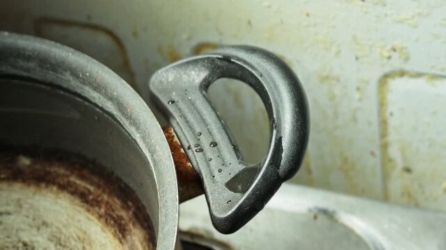 Close-up of a black pot handle with water droplets on worn metal cookware beside a stained kitchen sink, showing used household cookware in an indoor kitchen setting after cleaning.