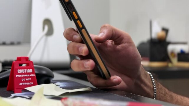 Close-up of a person using a smartphone at a desk with sticky notes and a humorous caution sign in a modern indoor office, captured with shallow focus and soft natural light.