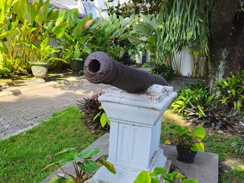 Historic cannon displayed on a stone pedestal at Keraton Yogyakarta courtyard, featuring metal barrel details, surrounding plants, paved ground, and traditional palace building in background.
