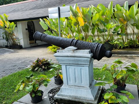 Historic cannon displayed on a stone pedestal at Keraton Yogyakarta courtyard, featuring metal barrel details, surrounding plants, paved ground, and traditional palace building in background.