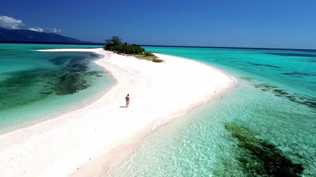Drone shot of a person on a pristine tropical shoreline and crystal-clear sea against a distant mountain range.