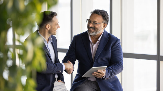 Two smiling businessmen sit together in office lounge, expressing mutual respect and shaking hands during meeting while holding digital tablet. Agreement, partnership building, business relationships