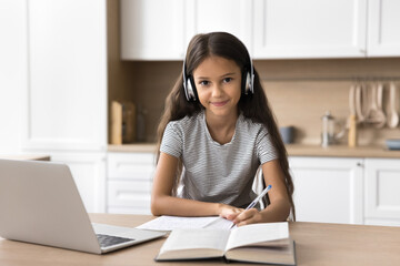 Portrait of cute little 7s schoolgirl studying at home using laptop, wear headphones listening audio course, improve english knowledge via application or educational platform. Modern tech, e-learning © fizkes