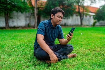 Obraz premium A man is sitting cross legged on a grassy lawn, holding a smartphone and seemingly engaged in a video call. The background features trees and a wall, suggesting an outdoor setting