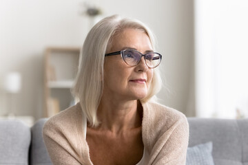 Beautiful pensive middle-aged woman wear glasses sit alone on couch and looking into distance with...