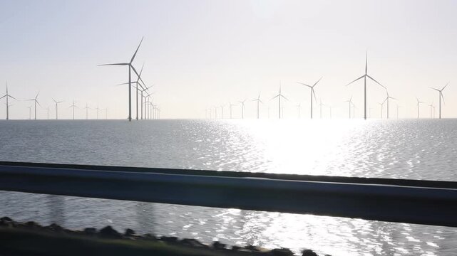 Low angle moving view with a blurred guardrail and offshore turbines in warm sunrise light.