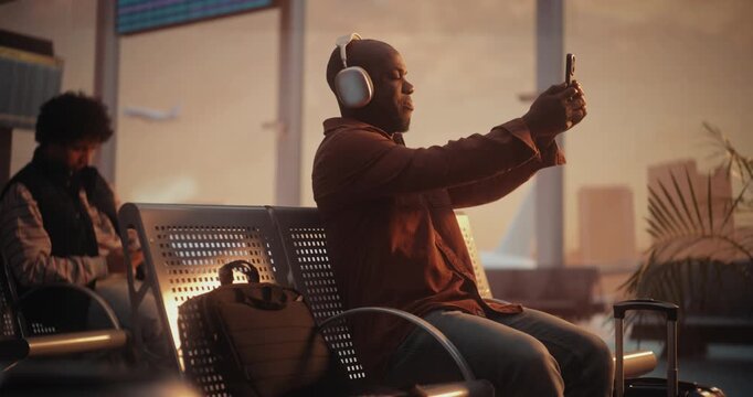 African American Man Sits in Airport Waiting Area and Captures Moment. Male Holds Smartphone Up With One Hand to Take Selfie. Staying Connected With Friends or Social Media During Flight Layover.