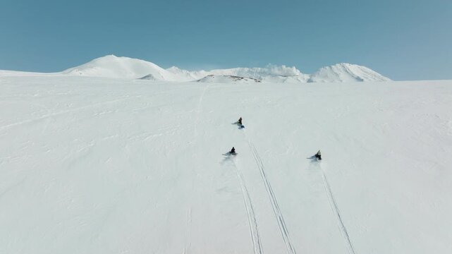 Snowmobilers riding across vast untouched snow slope leaving tracks on bright winter mountain, aerial rearview tracking ascends to reveal ridge