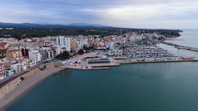 Delta de L&rsquo;Ebre, L&rsquo;Ampolla,Catalonia high drone reveal from marina , town and bay with sun reflecting off calm sea, mountains beyond and moody sky in bright late sunlight