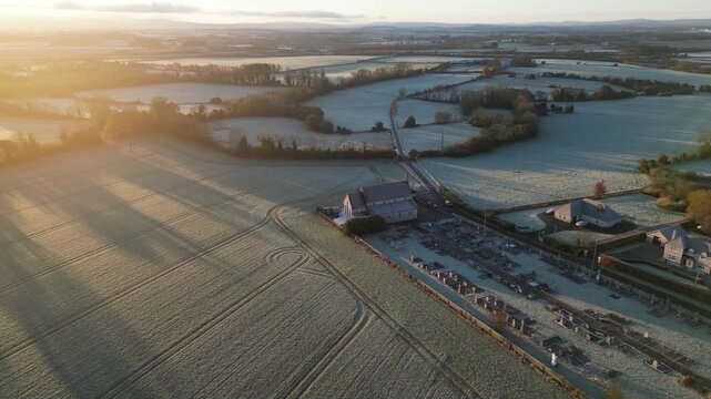 Breathtaking aerial footage of Saint Brendan's Church, Muckalee, Kilkenny, showcasing a historic cemetery surrounded by frost-kissed fields at sunrise. Crisp, serene winter landscape view.