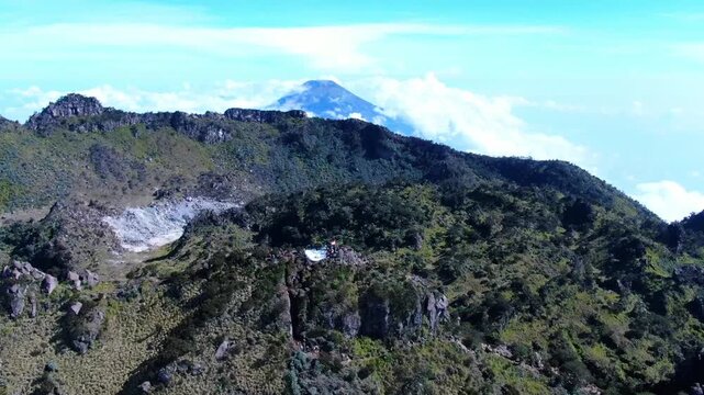 Cinematic dolly out drone shot reveals the rugged Puncak Sejati of Mount Sumbing, showcasing hikers gathered at the summit under a clear blue sky